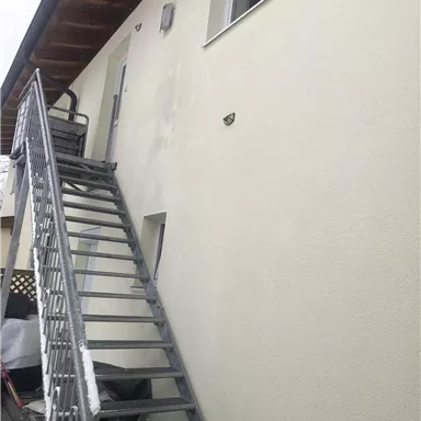 A gray, metallic staircase leads up to an entrance on a light wall. Above the stairs, there are two wall sconces.