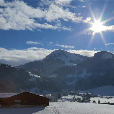 A snow-covered landscape with mountains in the background and bright sunshine. A red building stands in the foreground.