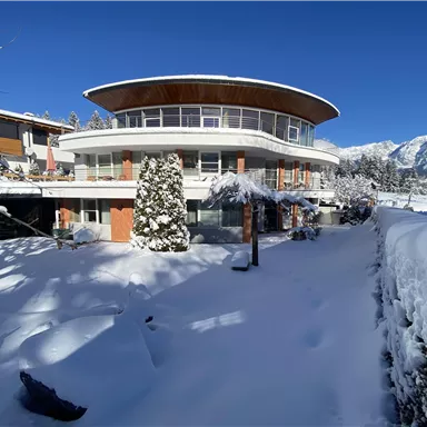 Ein modernes Haus in einer schneebedeckten Landschaft. Die Umgebung ist von Bergen und klarem blauen Himmel umgeben.
