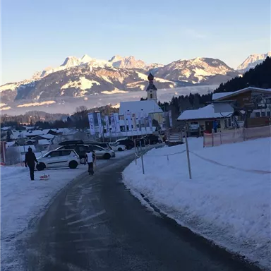 Eine schneebedeckte Landschaft mit Bergen im Hintergrund. Im Vordergrund führt eine Straße durch das winterliche Dorf mit Autos und Menschen.