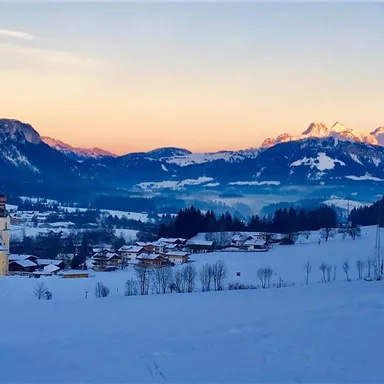 Eine verschneite Berglandschaft mit einem kleinen Dorf im Vordergrund. Im Hintergrund sind die schneebedeckten Berge und ein sanfter Sonnenaufgang zu sehen.