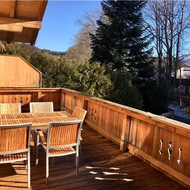 A cozy outdoor area with a wooden terrace and dining group. Surrounded by trees and clear sky.