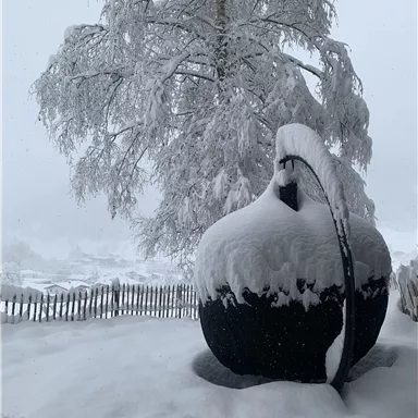 A snowy landscape with a large, snow-covered object and a tree in the background. The ground is completely covered with snow.