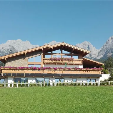 Ein malerisches Haus mit einem großen Balkon und bunten Blumen. Im Hintergrund erheben sich beeindruckende Berge unter einem klaren blauen Himmel.
