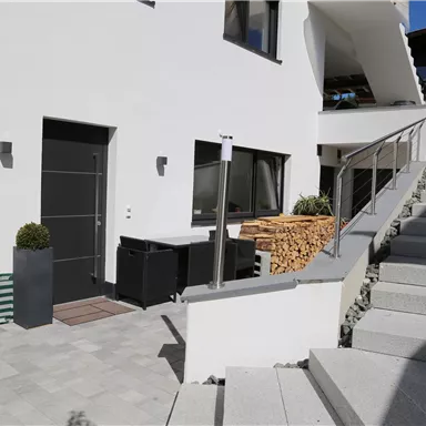 A modern house with a clean, white exterior and an inviting front door. The stone stairs lead to a well-maintained outdoor area with a wood store and plants.