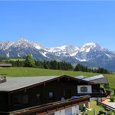 A beautiful mountain landscape with snow-covered peaks and a clear blue sky. In the foreground, traditional cottages can be seen in a green setting.
