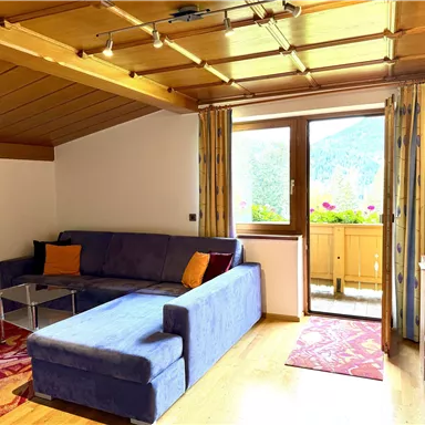 A cozy living room with a blue sofa and a window leading to a balcony with a view of flowers. The wooden ceiling and the carpet give the room a warm atmosphere.