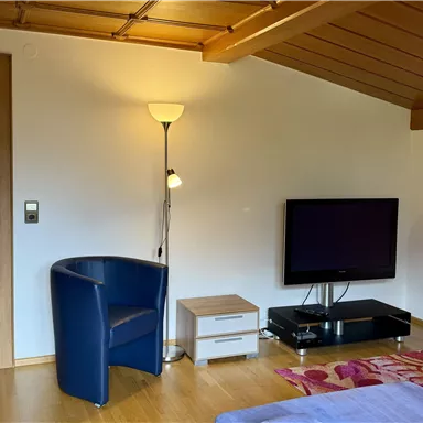 A cozy living space with a blue armchair, a television, and a small table. The walls are light, and the lighting creates an inviting atmosphere.