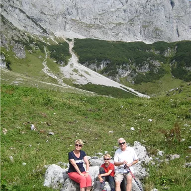 A family is sitting on a rock in nature. In the background, there are large mountains and a green meadow visible.