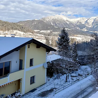 A charming house in a snowy landscape. In the background, majestic mountains and a blue sky are visible.
