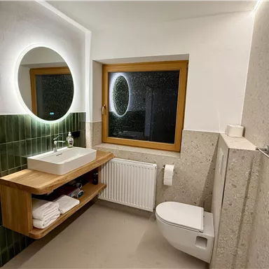A modern bathroom with a large mirror and a window. The walls are partially covered with green tiles, and there are wooden shelves with towels.