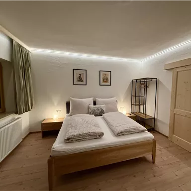 A modern bedroom with a double bed, two pillows, and a neutral color palette. Two pictures hang on the wall, and a metal shelf stands in the room.