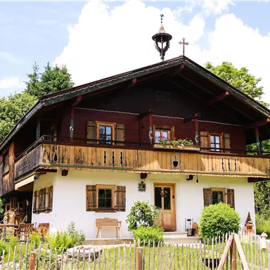 Ein charmantes altes Haus mit einem Holzbalkon und blühenden Fenstern. Es steht in einer grünen Umgebung mit Bäumen und Bergen im Hintergrund.