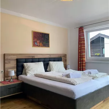 Ein gemütliches Schlafzimmer mit einem großen Bett und hellen Wänden. Neben dem Bett steht ein Nachttisch und ein Fenster sorgt für viel Tageslicht.