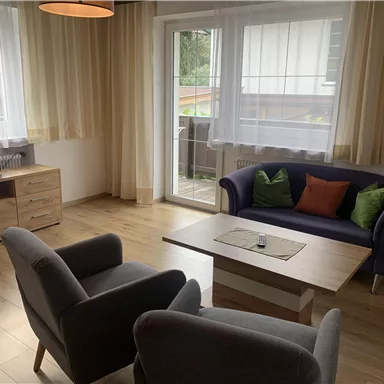 Ein gemütliches Wohnzimmer mit einem blauen Sofa und zwei grauen Sesseln. Große Fenster sorgen für viel Tageslicht.