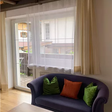 Ein gemütliches Wohnzimmer mit einem lila Sofa und bunten Kissen. Große Fenster lassen viel Tageslicht herein und bieten einen Blick auf einen Balkon.