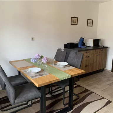 Eine gemütliche Küche mit einem Esstisch und vier Stühlen. Auf dem Tisch sind Teller, Gläser und frische Blumen dekoriert.