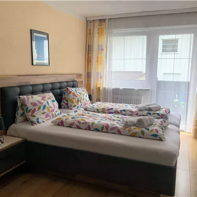 Ein gemütliches Schlafzimmer mit einem Doppelbett und bunten Bettbezügen. Große Fenster lassen viel Tageslicht herein.