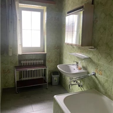 Ein Badezimmer mit grünen Fliesen und einer Badewanne. Das Fenster lässt viel Tageslicht herein.