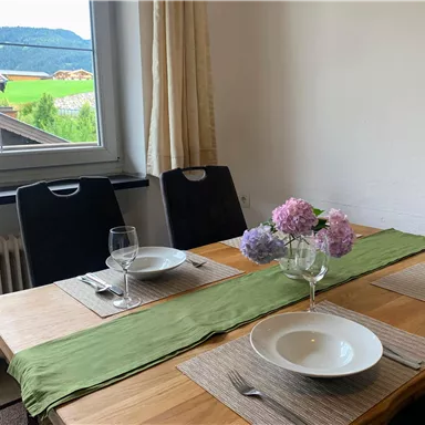 Ein gemütlicher Esstisch mit zwei gedeckten Tellern und einem Blumenstrauß. Der Blick aus dem Fenster zeigt eine grüne Landschaft und Berge im Hintergrund.