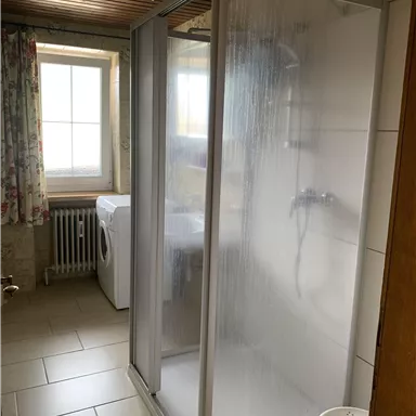 Ein Badezimmer mit einer Duschnische und einem Fenster. Im Hintergrund steht eine Waschmaschine und ein Hocker.