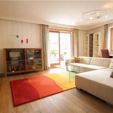 A modern living room with a white sofa and a large glass table. The room has large windows that let in plenty of natural light and a colorful rug on the wooden floor.
