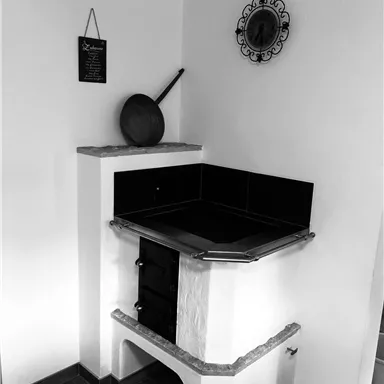 Ein traditioneller Holzofen in einer Ecke eines Raumes. Die Wände sind weiß und es gibt einige dekorative Elemente wie eine Pfanne und eine Wanduhr.