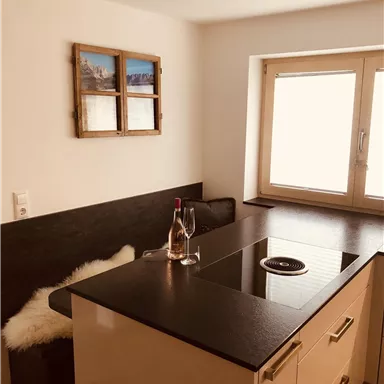 Eine moderne Küche mit einem eleganten schwarzen Kochfeld und einer gemütlichen Sitzecke. Große Fenster lassen viel Tageslicht herein.