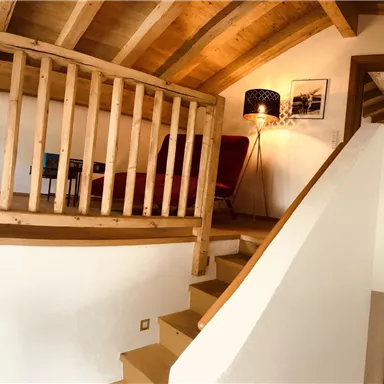 Ein modernes, helles Treppenhaus mit Holzbalustrade und warmem Licht. Im Hintergrund sind Stühle und Bilder erkennbar.