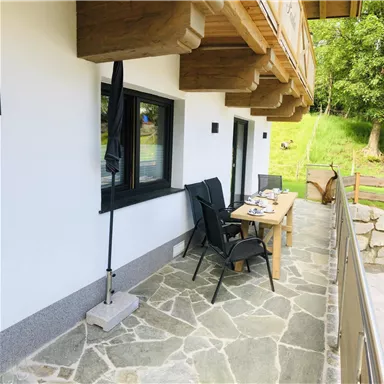 Ein gemütlicher Außenbereich mit einem Tisch und Stühlen auf einem steinernen Boden. Im Hintergrund ist eine grüne Wiese und ein Baum sichtbar.