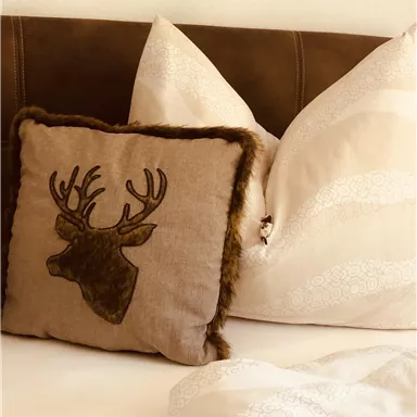 Ein gemütliches Bett mit flauschigen Kopfkissen und einem dekorativen Kissen mit einem Hirschmotiv. Die Farben sind warm und einladend.