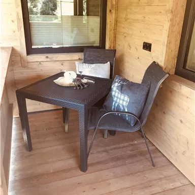 Ein gemütlicher Balkon mit einem Tisch und zwei Stühlen. Auf dem Tisch liegt eine kleine Dekoration und ein Kissen.