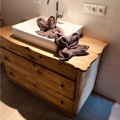 Ein elegantes Waschbecken aus weißem Keramik auf einem rustikalen Holzmöbel. Zwei handtuchartige Faltblumen liegen dekorativ auf der Ablage.