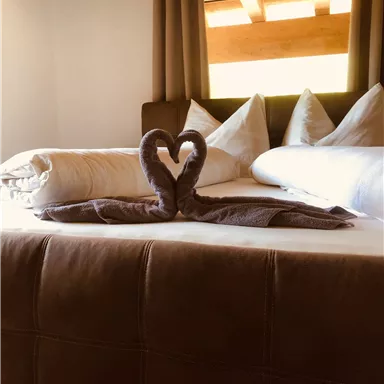 Ein gemütliches Schlafzimmer mit einem großen Bett und dekorativen Kissen. Ein Handtuch in Herzform liegt in der Mitte des Bettes.