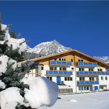 Ein modernes Gebäude im Schnee, umgeben von Bergen. Im Vordergrund steht ein Baum mit Schnee auf den Zweigen.