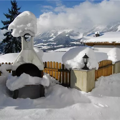 A snowy landscape with a grill and lanterns covered in snow. In the background, mountains and a blue sky can be seen.