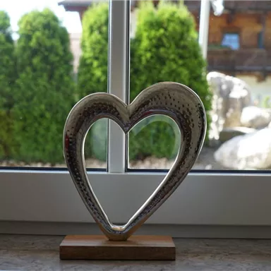 A decorative heart sculpture made of silver stands on a wooden base. In the background, you can see green trees and a part of a house.