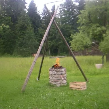Ein gemütliches Lagerfeuer auf einem Stehständer in einer grünen Wiese. Im Hintergrund sind Bäume und ein Huhn zu sehen.