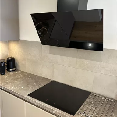 Eine moderne Küche mit einem schwarzen Cerankochfeld und einer eleganten Dunstabzugshaube. Der Arbeitsbereich ist in neutralen Farbtönen gehalten und hat einen klaren, minimalistischen Look.