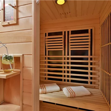 Eine gemütliche Sauna mit Holzverkleidung und zwei Handtüchern. Neben der Sauna stehen zwei Gläser mit Getränken auf einem kleinen Tisch.