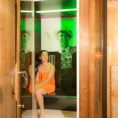 A woman in an orange towel sits in a modern sauna with green lighting. In the background, there is a depiction of a person.