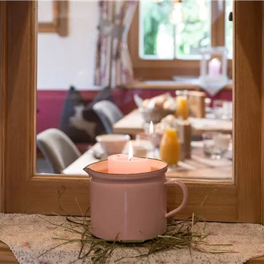 Ein gemütlicher Blick durch ein Fenster auf einen gedeckten Tisch. Eine brennende Kerze in einer rosa Tasse steht im Vordergrund.