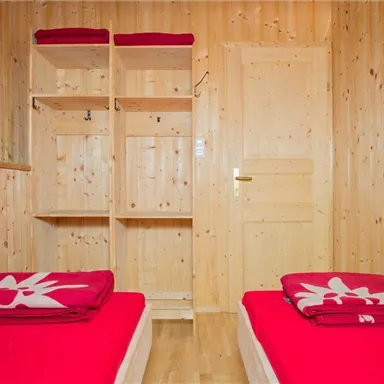 Ein helles Zimmer mit Holzverkleidung und zwei Einzelbetten. Rote Bettwäsche und ein offenes Regal vervollständigen den Raum.
