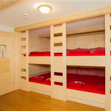 Ein Zimmer mit zwei Stockbetten aus hellem Holz und roten Bettdecken. Der Raum hat eine einfache, saubere Einrichtung und einen warmen Holzfußboden.