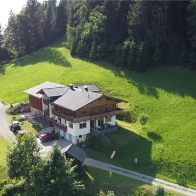 Ein gemütliches Haus auf einer grünen Wiese, umgeben von Bäumen und Bergen. Im Vordergrund gibt es einen Platz für sportliche Aktivitäten.