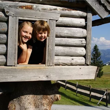 Zwei Kinder schauen aus einem Fenster eines Holzhäuschens. Im Hintergrund sind grüne Hügel und ein blauer Himmel zu sehen.