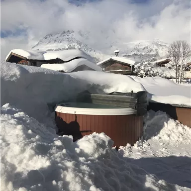 A snowy garden with a hot tub and snow underneath. In the background, mountains and a blue sky can be seen.