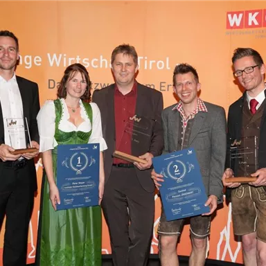 Eine Gruppe von vier Personen steht auf einer Bühne. Sie halten Auszeichnungen und Plaketten in der Hand, während im Hintergrund ein orangefarbener Banner von WKO zu sehen ist.