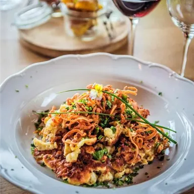Ein Teller mit köstlichen Röstkartoffeln, garniert mit frischem Schnittlauch und knusprigen Zwiebeln. Im Hintergrund sind ein Glas Rotwein und mehrere kleine Gerichte zu sehen.