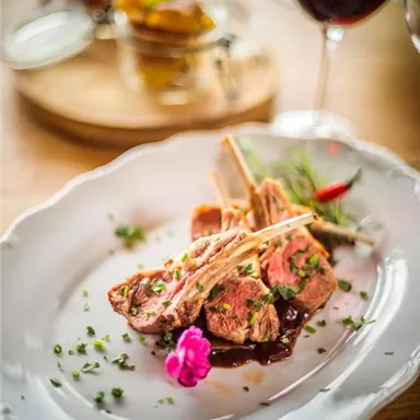 Ein köstliches Gericht mit Lammkoteletts, garniert mit einer Blume und Kräutern. Im Hintergrund sind ein Glas Rotwein und eine kleine Schale mit Beilagen zu sehen.